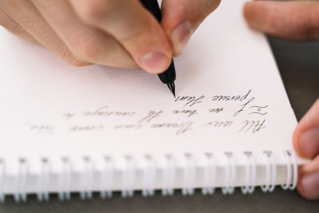 A person writes in a spiral notebook with elegant handwriting, using a pen.